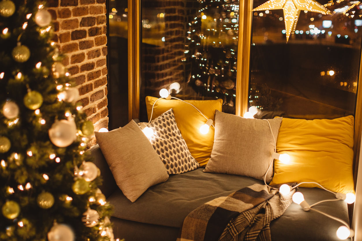 Artigos de Natal: à esquerda há uma árvore de Natal desfocada enfeitada com bolas e luzes brancas. Ao lado, um sofá focado com almofadas coloridas e luzes sobre seu encosto.