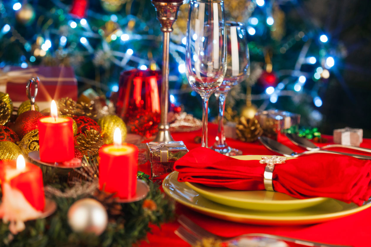 Artigos de Natal: mesa da Ceia decorada com velas vermelhas, festão verde, bolas prateadas, vermelhas e douradas, além de um guardanapo de pano vermelho. Ao fundo, luzes brancas desfocadas.