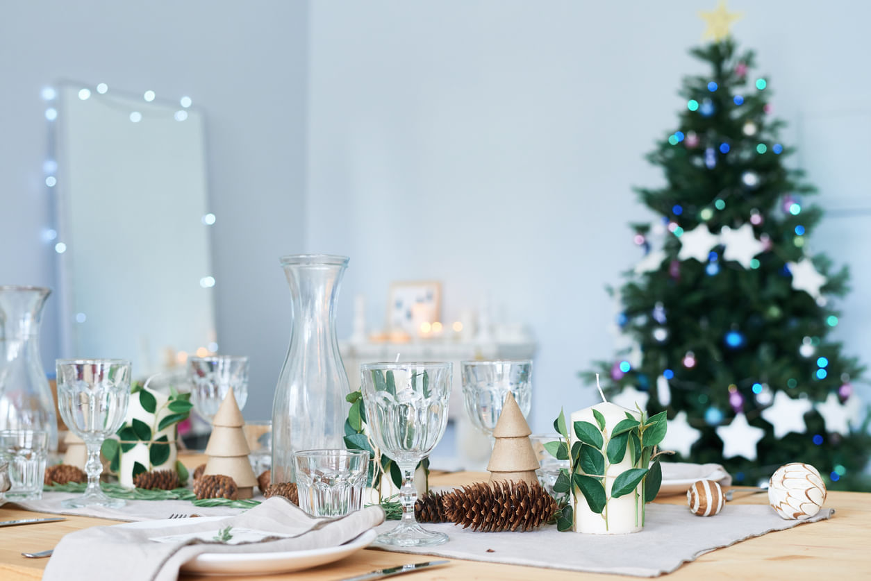Artigos de Natal: mesa de jantar com copos e jarras transparentes, velas brancas envoltas com folhas, bolas enfeitadas e enfeites com a silhueta de uma árvore de Natal. Ao fundo, uma parede branca e uma árvore de Natal decorada com luzes coloridas e estrelas brancas