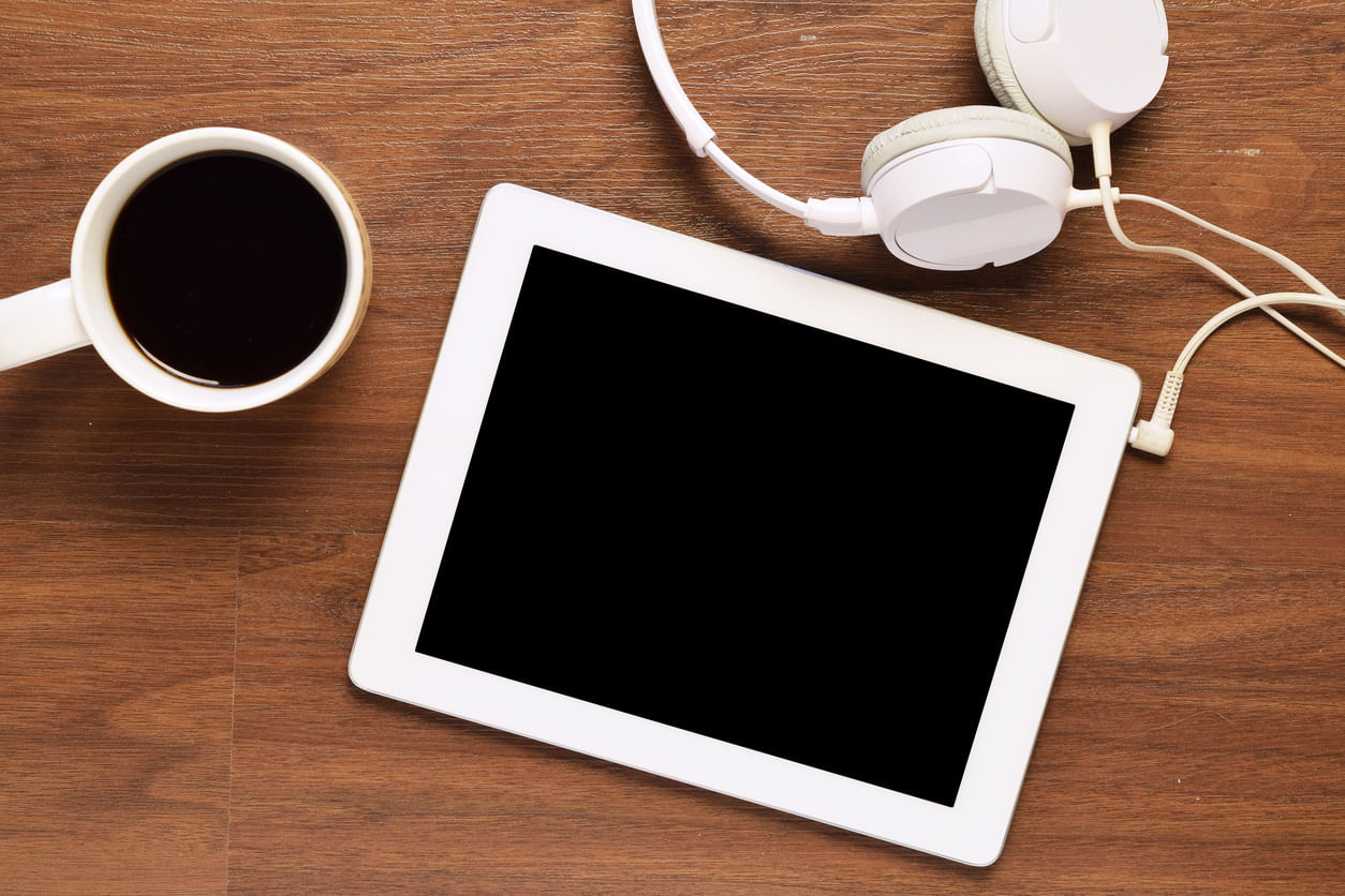 Promoções Black Friday: da esquerda para a direita temos uma xícara com café, um tablet branco e, acoplado a ele, um headphone. Vistos de cima, todos esses objetos são brancos e estão sob uma superfície de madeira.