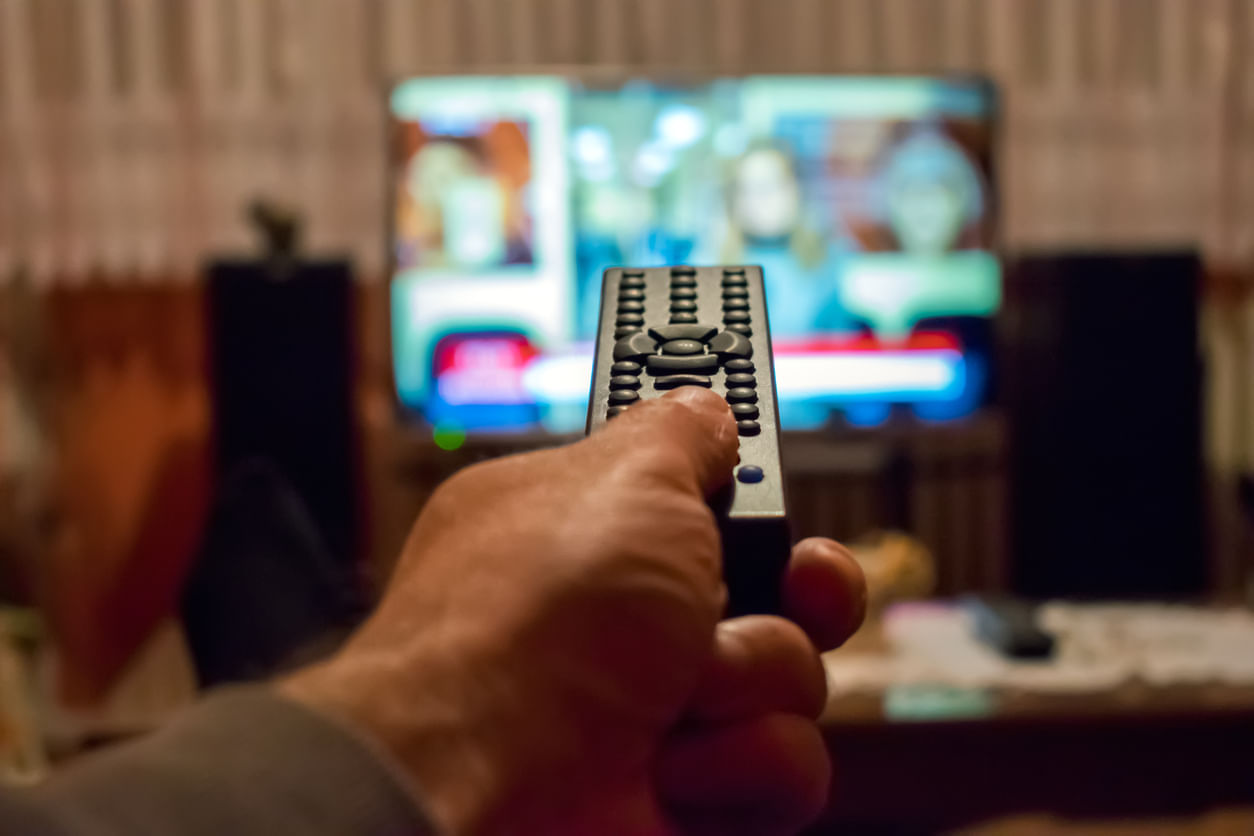 Promoções Black Friday: No primeiro plano, aparece a mão de uma pessoa segurando o controle remoto apontado para a TV, que está desfocada no fundo da imagem.