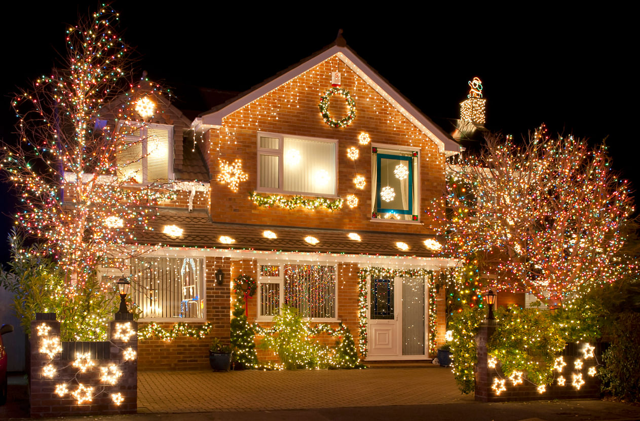 Enfeites de Natal: frente de um sobrado feito com tijolos toda enfeitada com luzes coloridas (árvores, arbustos, contornos das janelas e porta). Há uma guirlanda sobre a janela do andar de cima e um Papai Noel iluminado no topo da chaminé, do lado esquerdo do telhado.