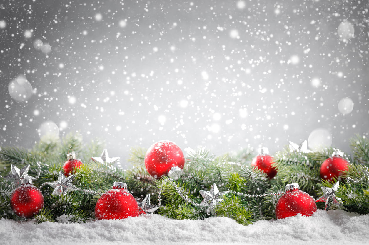 Enfeites de Natal: fundo branco com pontos de luz desfocados caindo como flocos de neve. À frente, galhos de árvores de Natal enfeitados com bolas vermelhas e estrelas prateadas