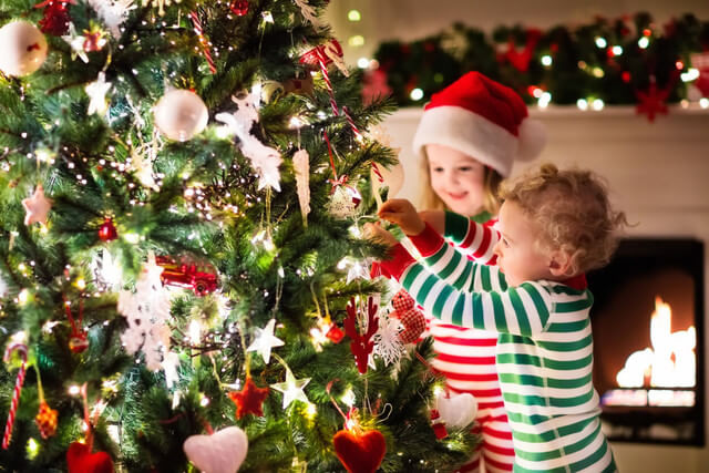 Ofertas de Natal 2018: duas crianças pequenas, com pijamas listrados, montam uma árvore de Natal com bolas, estrelas e pisca-pisca. Ao fundo, há enfeites natalinos e uma lareira